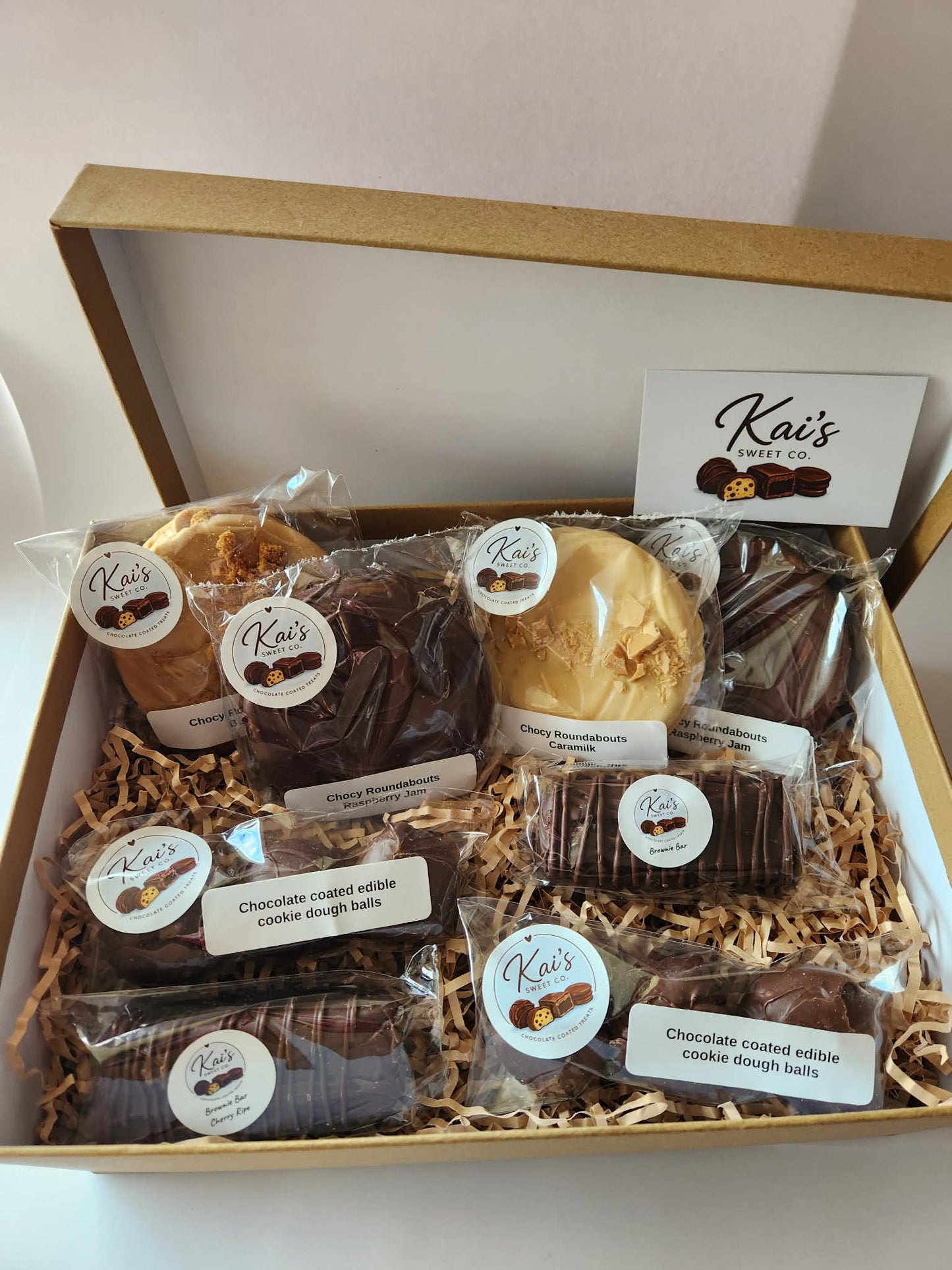 Kai's Sweet Co Large Gift Box open showing four chocolate roundabouts, cookie dough balls and two brownie bars nestled in shredded paper in a kraft box with Kai's Sweet Co gift card